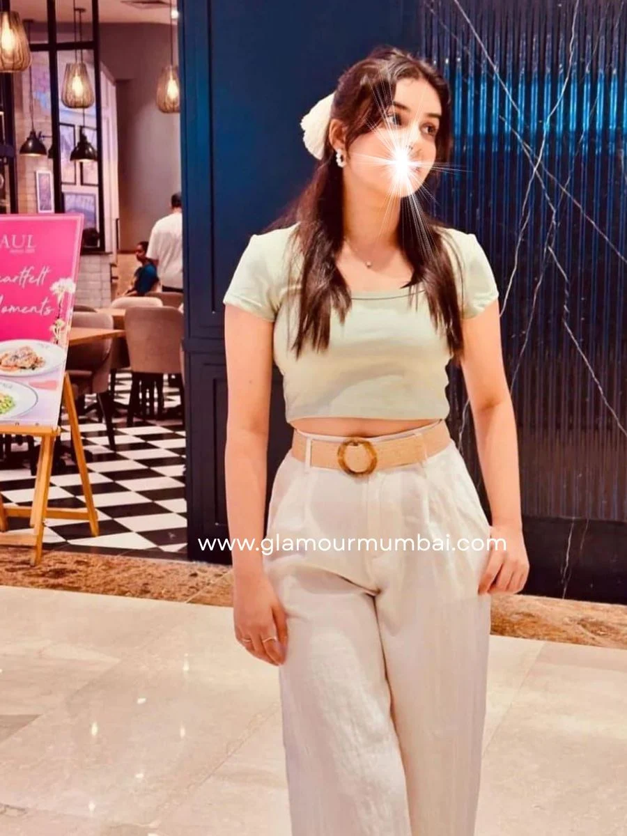 Pallavi escort profile photo indoors in Mumbai, standing near patterned wall with furniture in background, looking at camera.